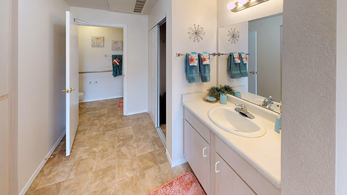 A clean bathroom interior featuring a white sink with a countertop, a large mirror above it, and a set of teal and floral hand towels hanging on a towel rack. The floor has beige tiles, and there is a partially open door leading to a toilet area with more teal towels and wall art. The walls are painted white, and there is a pink bath mat on the floor.