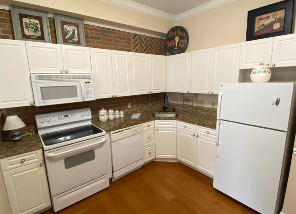 A kitchen with white cabinets, a white refrigerator, a white stove with an oven, a white microwave above the stove, a white dishwasher, and a granite countertop. The backsplash is a brick pattern, and there are decorative framed pictures and a round vintage sign on the walls. The floor is wooden.