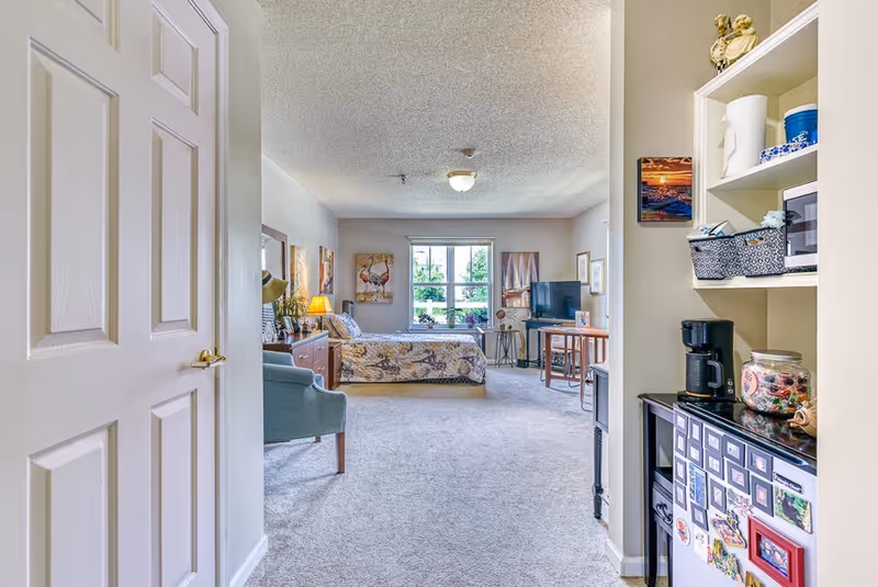 View into a senior living apartment showing a bedroom area with a bed, dresser, armchair, and TV. To the right, there is a small kitchenette area with a coffee maker, microwave, and mini fridge decorated with magnets. The room has a window letting in natural light and light-colored walls and carpet.