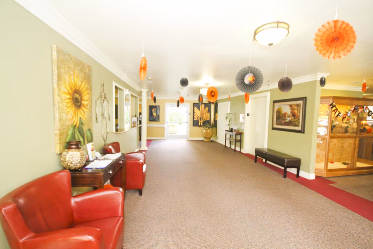 A wide hallway in an assisted living facility decorated with hanging orange and black paper decorations. The hallway has green walls adorned with framed artwork and a sunflower painting. There are red leather chairs and a wooden table with decorative items on the left side, and a black bench on the right. The far end of the hallway has a glass door letting in natural light.