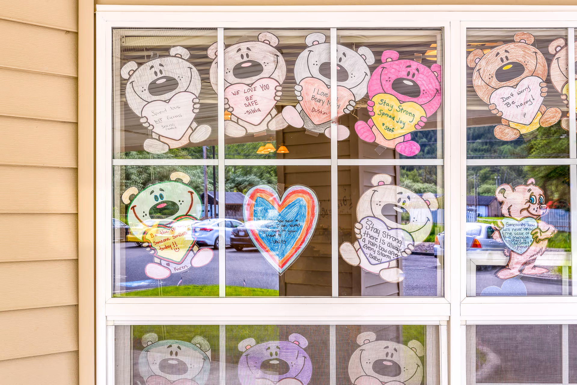 Window decorated with colorful paper cutouts of bears holding hearts with positive messages such as 'Stay Strong', 'We Love You Be Safe', and 'Don't Worry Be Happy'. A heart shape with a rainbow outline is also displayed in the center. The window reflects parked cars and greenery outside.