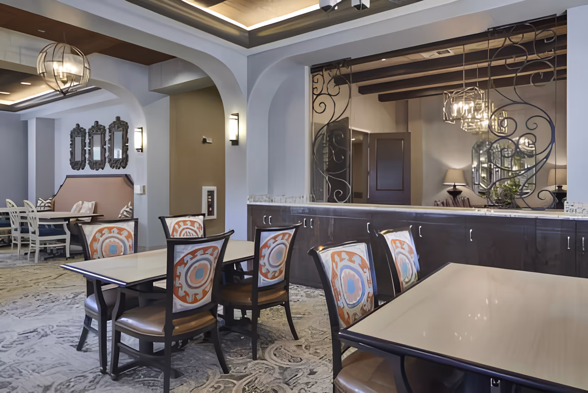 A dining area in an assisted living facility featuring tables with chairs that have colorful patterned backs. The room has decorative mirrors on the walls, wall sconces for lighting, and an ornate metal divider with a mirror behind it. The ceiling has recessed lighting and wooden beams, creating a warm and inviting atmosphere.