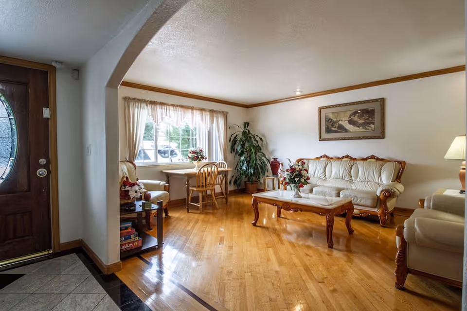 A cozy living room with wooden flooring, a cream-colored sofa set with wooden accents, a wooden coffee table with a floral arrangement, a small table with two chairs near a window with sheer curtains, a large potted plant in the corner, and a wooden front door with an oval glass panel.