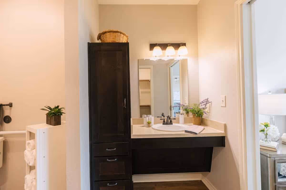 A bathroom vanity area with a dark wood cabinet and drawers on the left, a white countertop with a sink, a mirror above the sink, and three light fixtures mounted above the mirror. There are decorative plants on the countertop and a basket on top of the cabinet. To the left, a partial view of a toilet with a towel rack holding rolled white towels is visible. To the right, an open doorway leads to a bedroom with a bedside table, lamp, and flowers.