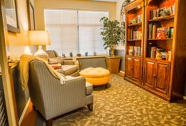 A cozy living room area with two upholstered armchairs, a round yellow ottoman, a wooden bookshelf filled with books and decorative items, a potted plant, a window with closed blinds, and a table lamp on a side table.