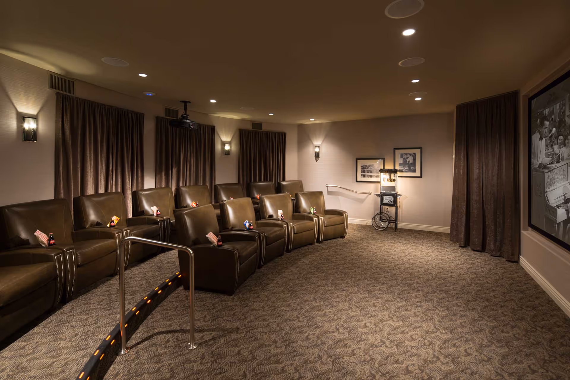 A cozy home theater room with two rows of brown leather recliner chairs, each with a small snack and drink placed on the armrest. The room has dim lighting, carpeted floor, wall sconces, and dark curtains covering the windows. A popcorn machine is visible in the back corner, and a black and white movie is playing on a large screen on the right wall.