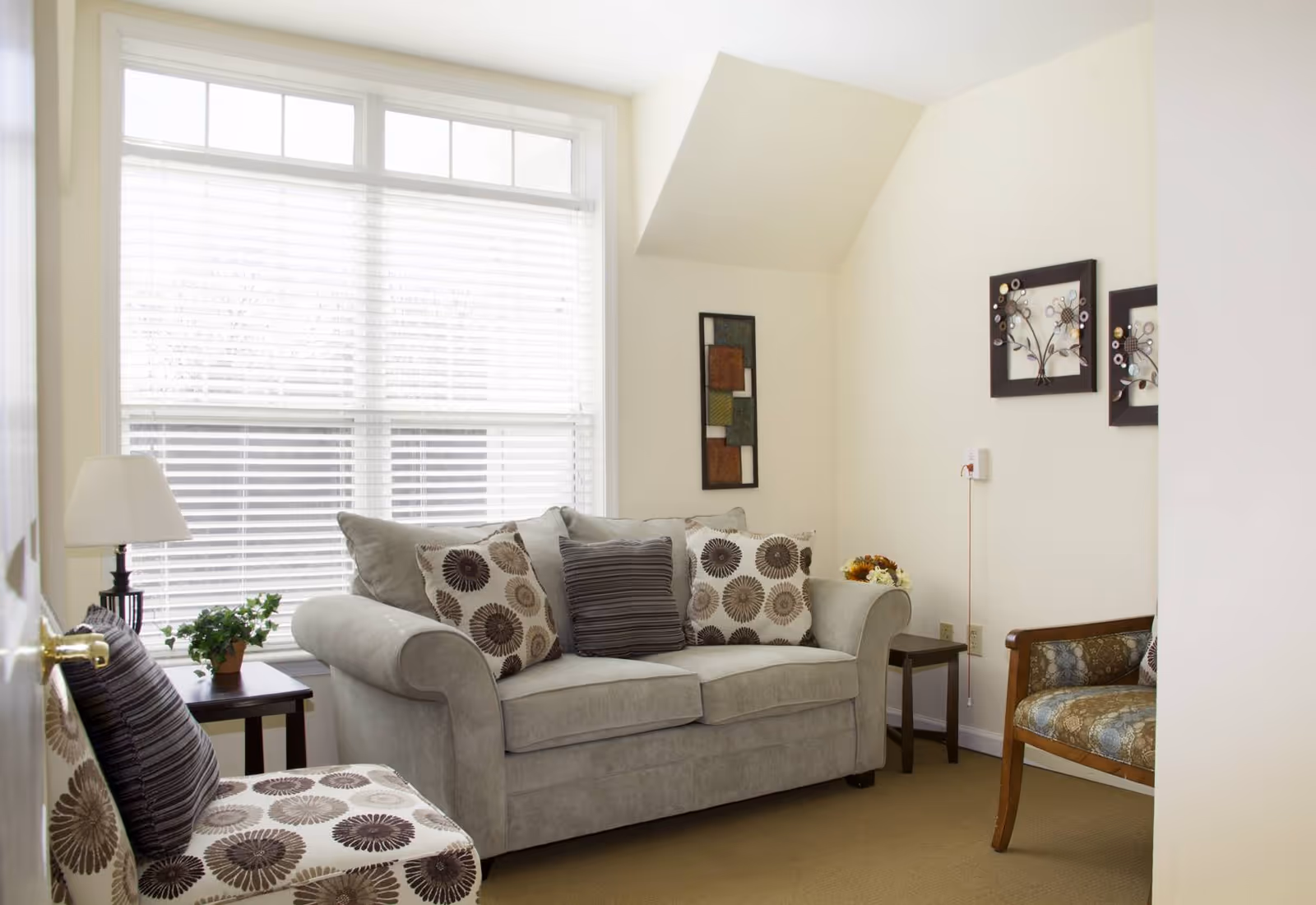 A cozy living room with a light gray sofa adorned with patterned and striped cushions, a matching patterned armchair, a wooden chair with floral upholstery, a small side table with a lamp and a potted plant, and wall decorations including framed art pieces. The room is well-lit by a large window with white blinds.