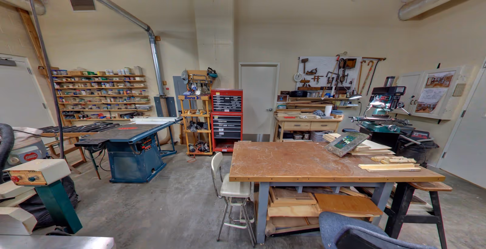 A workshop room with various woodworking tools and equipment including a table saw, workbenches, shelves with supplies, a red tool chest, and a drill press. The room has concrete floors and beige walls with tools hanging on a pegboard.
