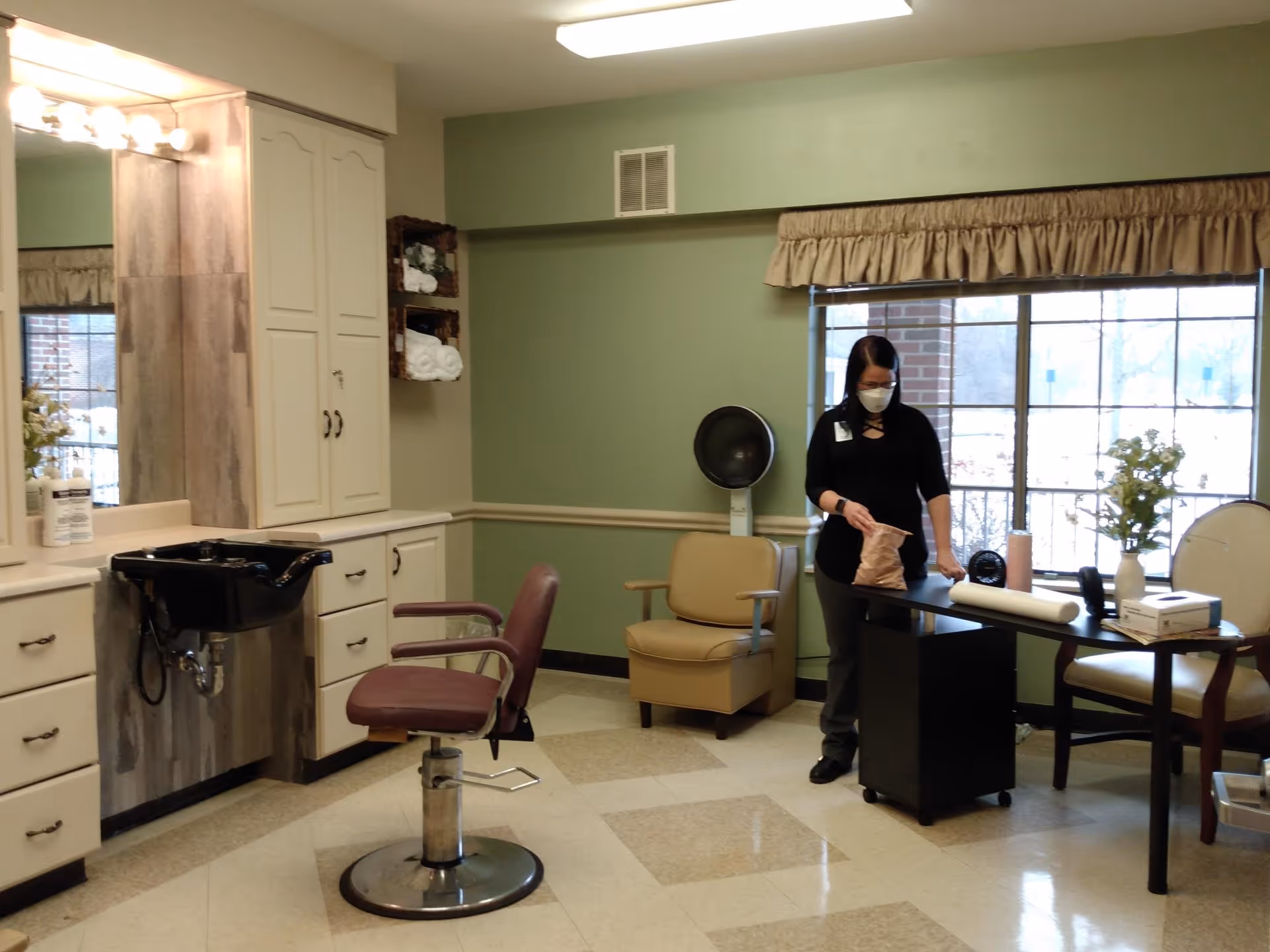 A hair salon area inside an assisted living facility with a salon chair and sink on the left, a woman wearing a mask standing near a table with various items, and a large window with beige curtains in the background.