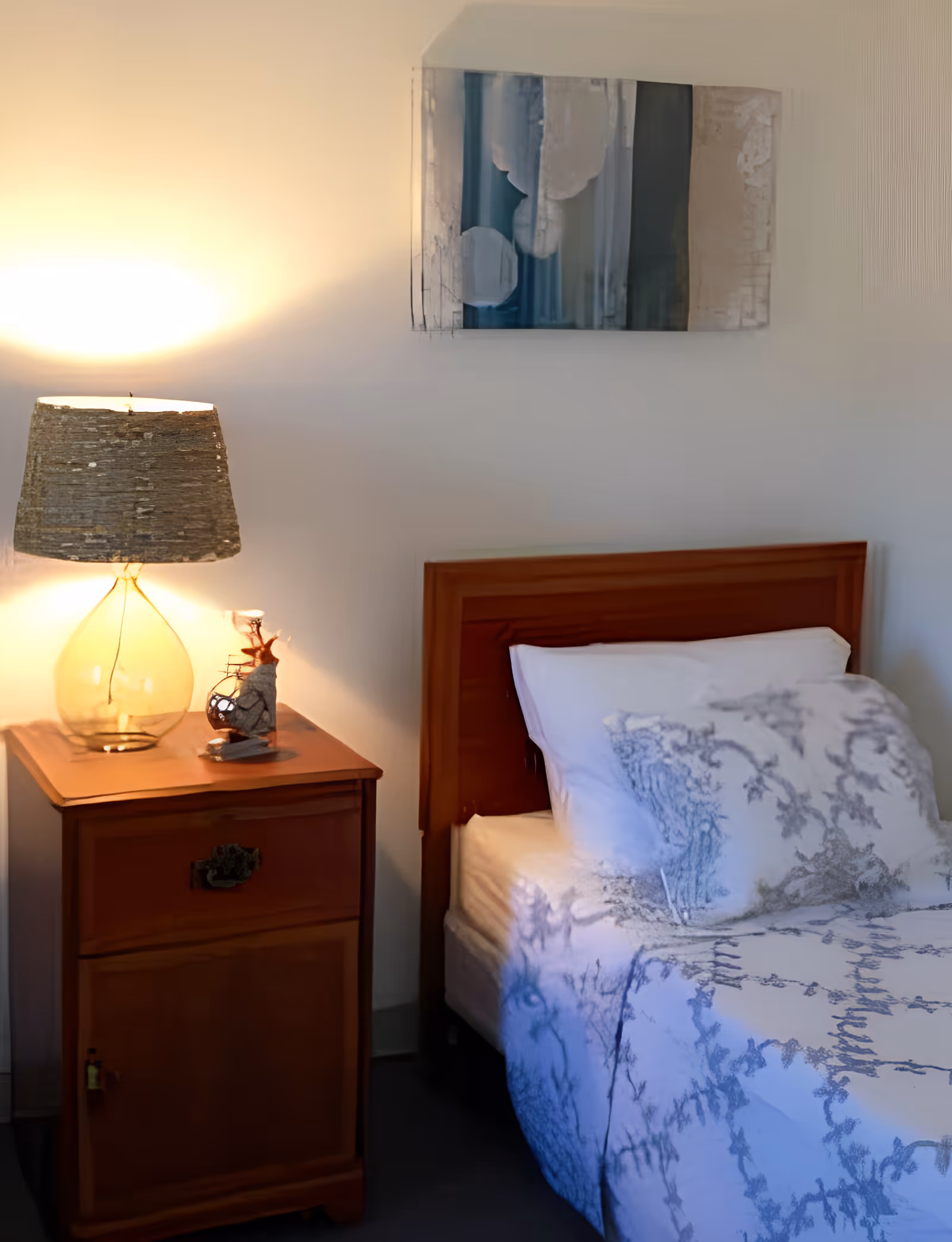A wooden nightstand with a glass lamp and decorative item beside a single bed with patterned bedding and a wooden headboard, under wall art.