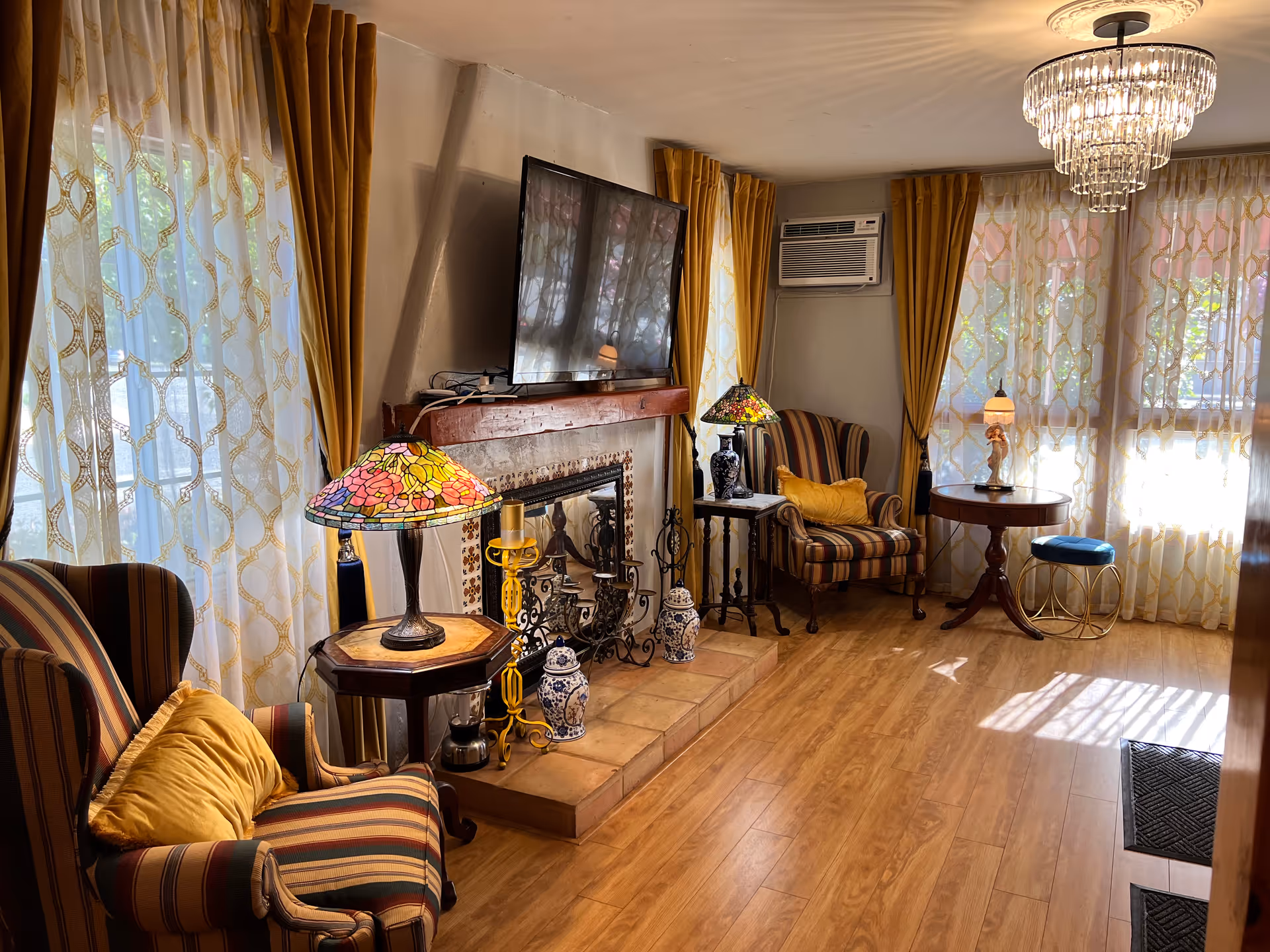 A cozy living room with wooden flooring, two striped armchairs with yellow cushions, a fireplace with decorative items, a flat-screen TV mounted above the fireplace, stained glass table lamps on small side tables, large windows with sheer patterned curtains and mustard yellow drapes, an air conditioning unit, a round wooden table with a small lamp, and a crystal chandelier hanging from the ceiling.