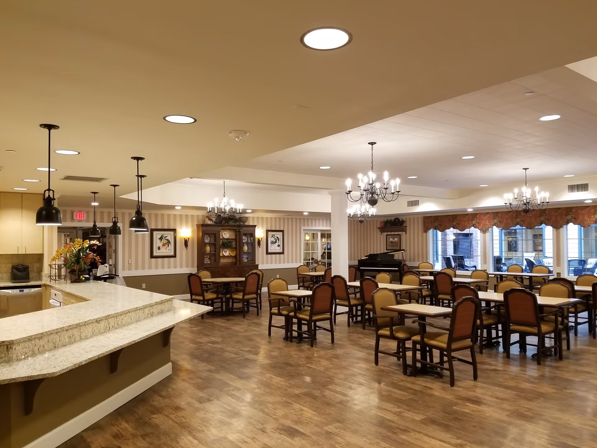 Spacious dining room with multiple tables and chairs arranged neatly. The room features a wooden floor, chandeliers hanging from the ceiling, large windows with floral valances, and a piano in the corner. A countertop with pendant lights is visible on the left side.