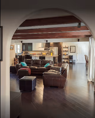 View through an arched doorway into a spacious living area with dark wooden floors and exposed wooden ceiling beams. The room features a brown leather sectional sofa with teal and red cushions, a matching ottoman, and a modern kitchen with stainless steel appliances in the background. There is a dining area visible to the right and shelves with decorative items near the back wall.