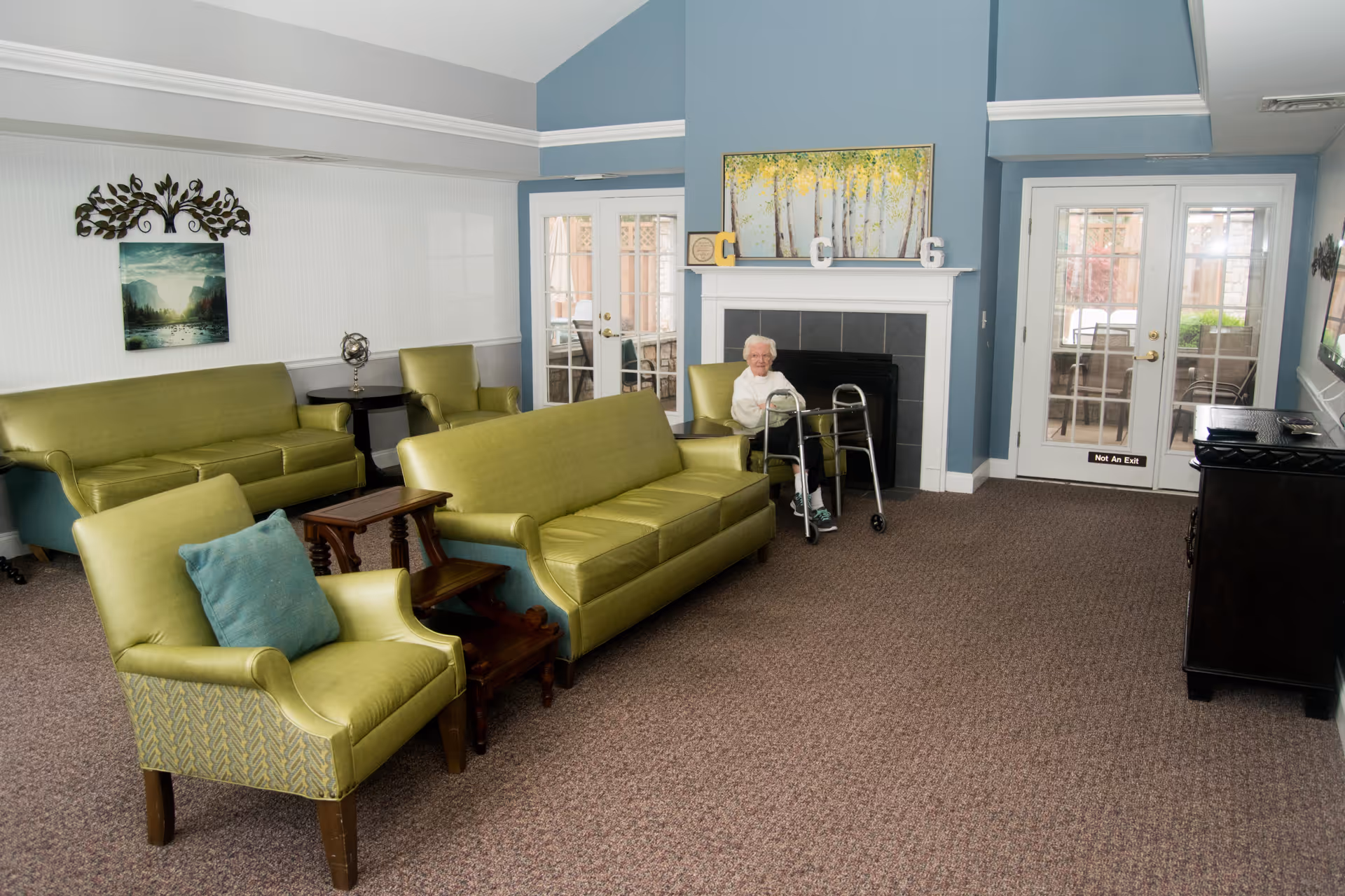 A senior living community lounge with green upholstered sofas and armchairs arranged around a fireplace. An elderly woman with a walker is seated near the fireplace. The room has blue and white walls, carpeted floor, and glass doors leading outside. There is a painting above the fireplace and decorative items on the mantel.