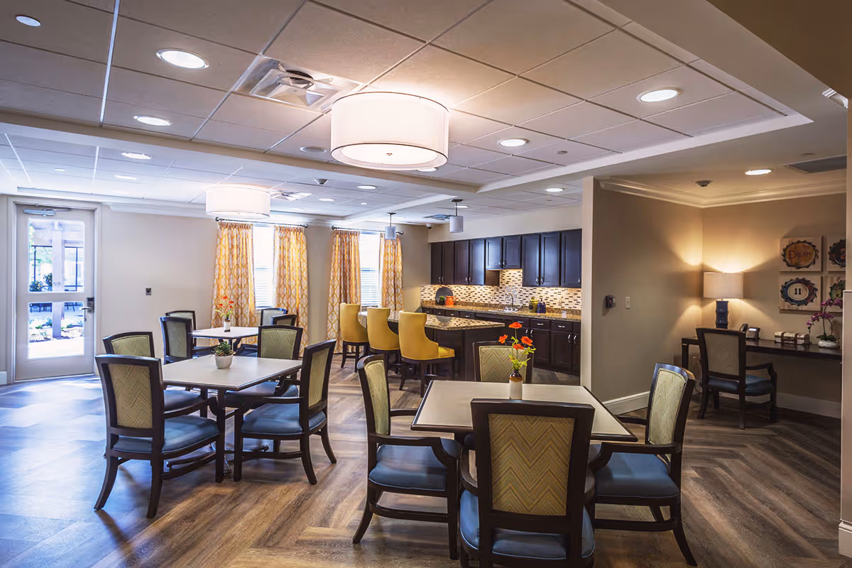 A bright and spacious dining area in a senior living facility featuring several square tables with chairs around them. The room has large windows with yellow patterned curtains, a kitchen area with dark wood cabinets and a tiled backsplash, and a counter with yellow bar stools. There is also a small desk with a lamp and decorative wall art in the corner.