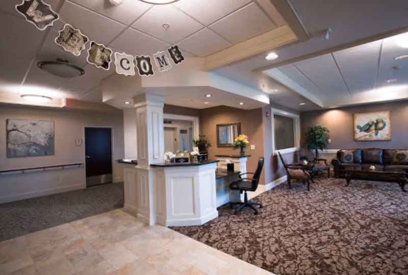 Interior view of a senior living facility reception area with a white reception desk, an office chair, and a seating area with a brown patterned carpet, a dark brown leather sofa, two chairs, a small table, and a painting of birds on the wall. A 'WELCOME' banner hangs from the ceiling.