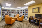 A cozy library or reading room with white bookshelves filled with books along the walls. Four orange armchairs and a round ottoman are arranged in the center on a patterned carpet. A wooden desk with a computer and chair is positioned against a yellow wall with a framed artwork above it. The ceiling has recessed lighting panels.