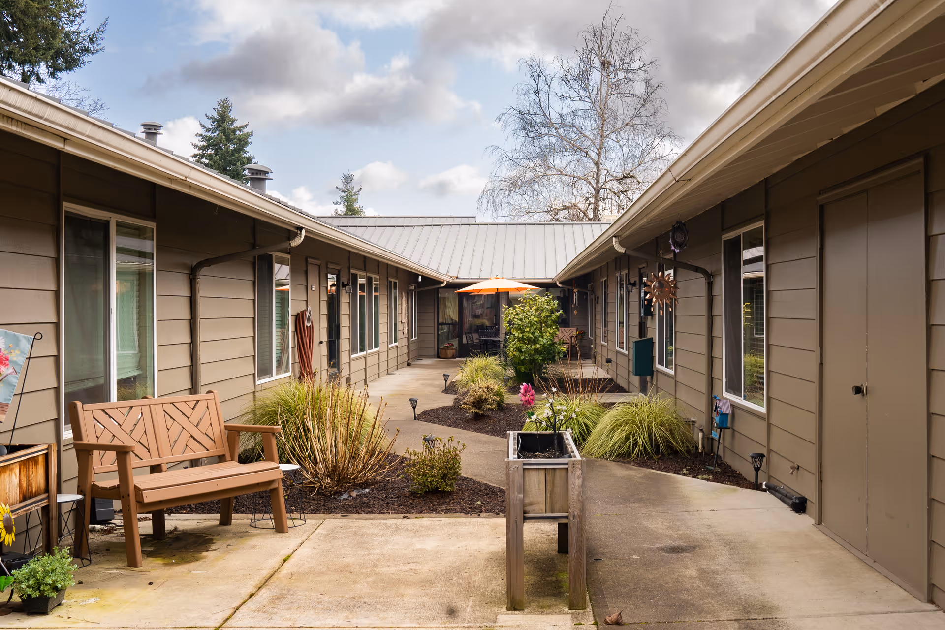 Outdoor courtyard area of Jefferson Lodge Memory Care Community featuring a concrete walkway, garden beds with shrubs and plants, a wooden bench, and an orange patio umbrella in the background. The courtyard is surrounded by single-story buildings with windows and doors.