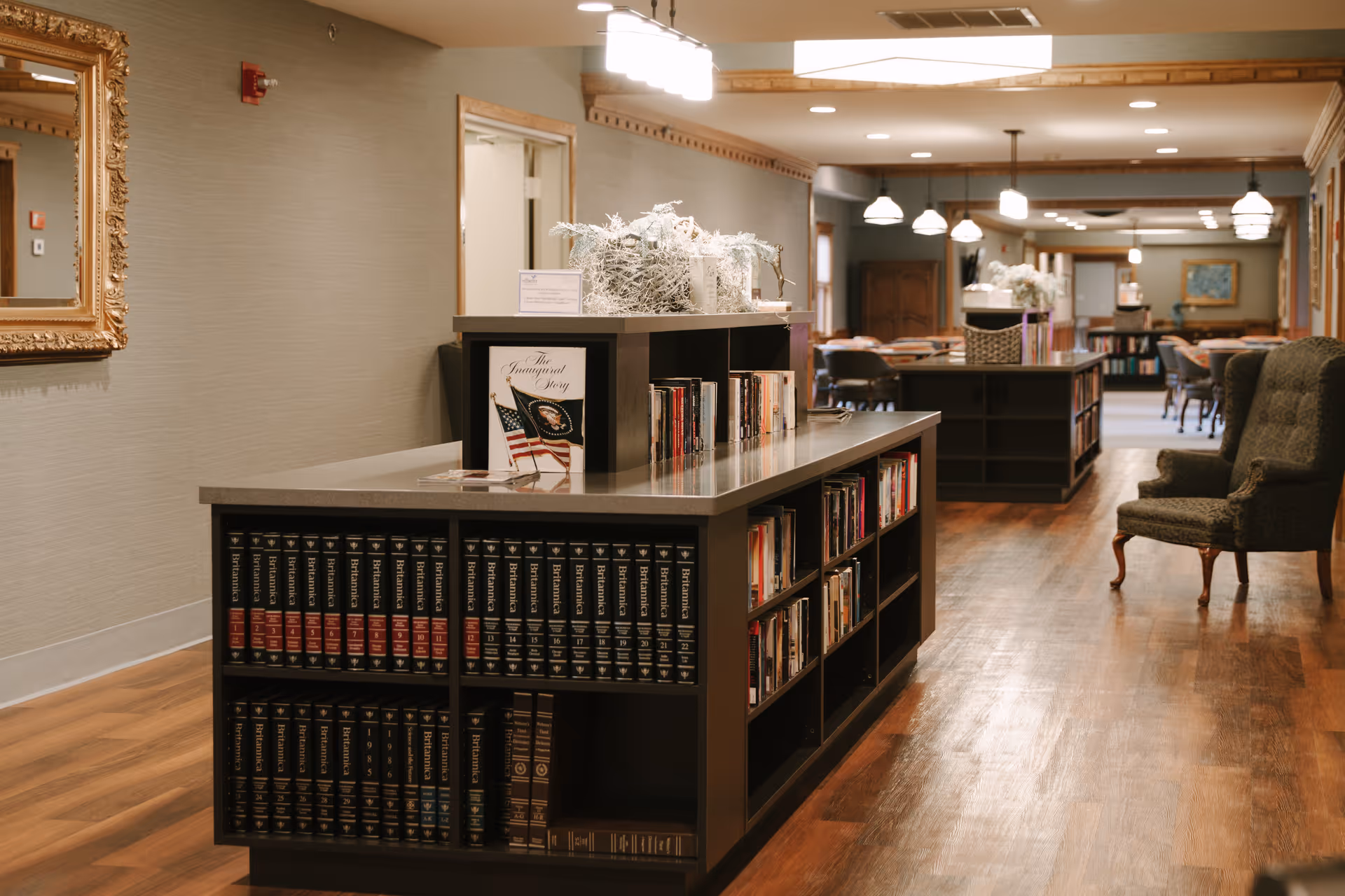 Interior view of a senior living facility hallway with wooden flooring and bookshelves filled with books. There are comfortable armchairs on the right side and decorative plants on top of the bookshelves. The hallway is well-lit with ceiling lights and has framed artwork and mirrors on the walls.