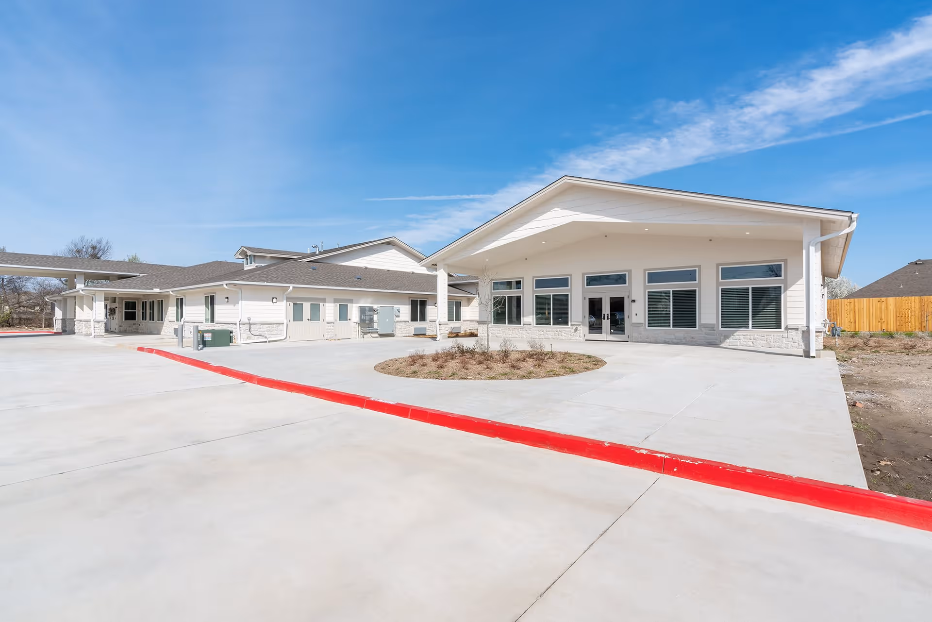 Exterior view of a single-story senior living facility building with a large concrete driveway and a circular landscaped area in front. The building has a modern design with white siding, large windows, and a covered entrance. The sky is clear and blue.