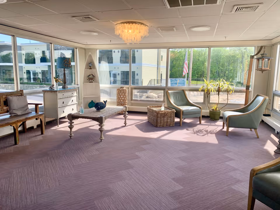 Sunlit common room with armchairs, a coffee table, and large windows overlooking the grounds.