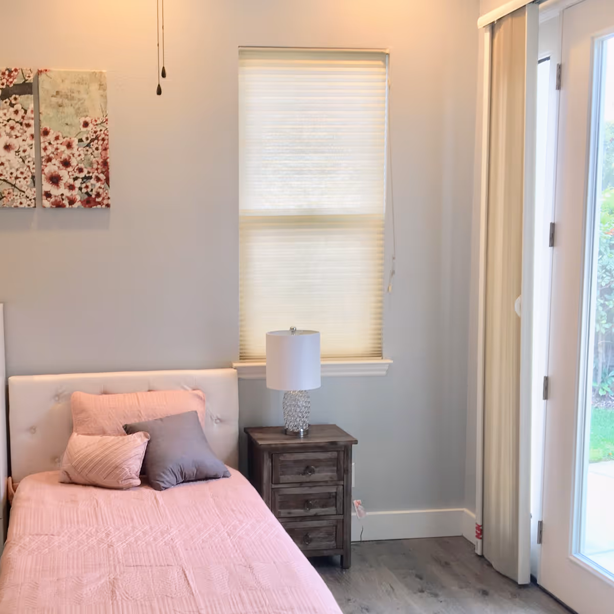A cozy bedroom with a single bed covered in a pink quilt and three pillows in shades of pink and gray. Next to the bed is a wooden nightstand with three drawers and a decorative glass lamp on top. A window with a closed blind is above the nightstand, and to the right, there is a glass door with vertical blinds partially open, showing a glimpse of greenery outside. Two floral paintings hang on the wall above the bed.