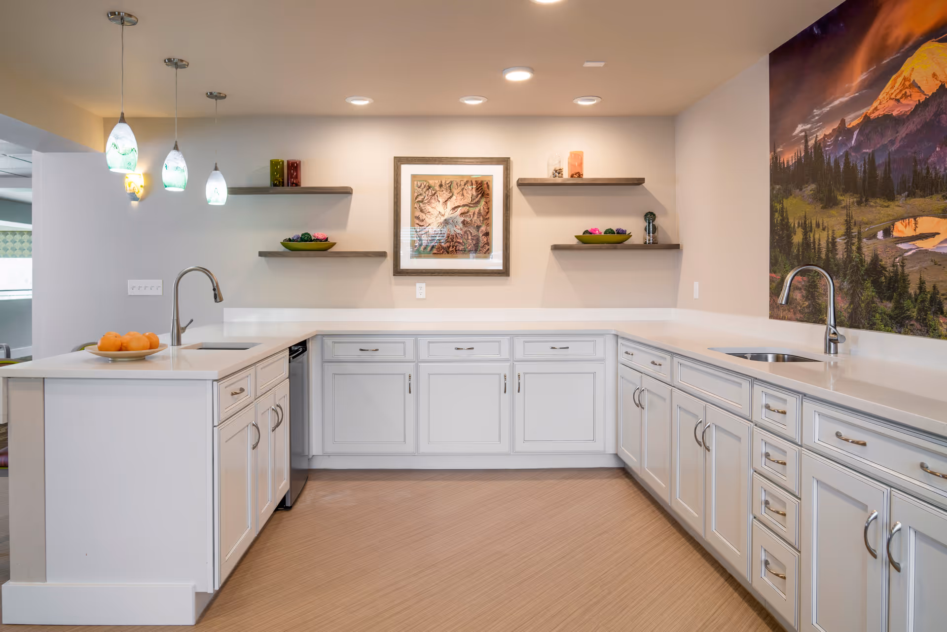 A bright, modern kitchen with white cabinets and countertops. There are two sinks with chrome faucets, three pendant lights hanging above the counter on the left, and three floating shelves on the back wall displaying decorative items. A large scenic painting of a mountain landscape is on the right wall.