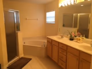 A bathroom with a corner bathtub, a glass-enclosed shower, a double sink vanity with wooden cabinets, a large mirror above the sinks, a small window, and beige tiled floor and walls.