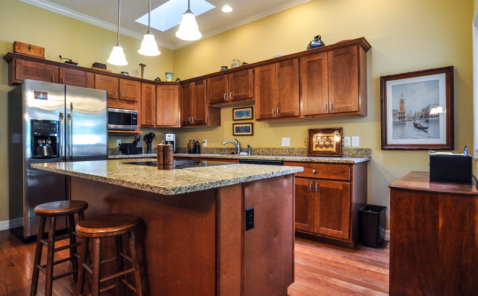 Kitchen with a large granite island, wooden cabinets, stainless steel refrigerator, and pendant lighting.