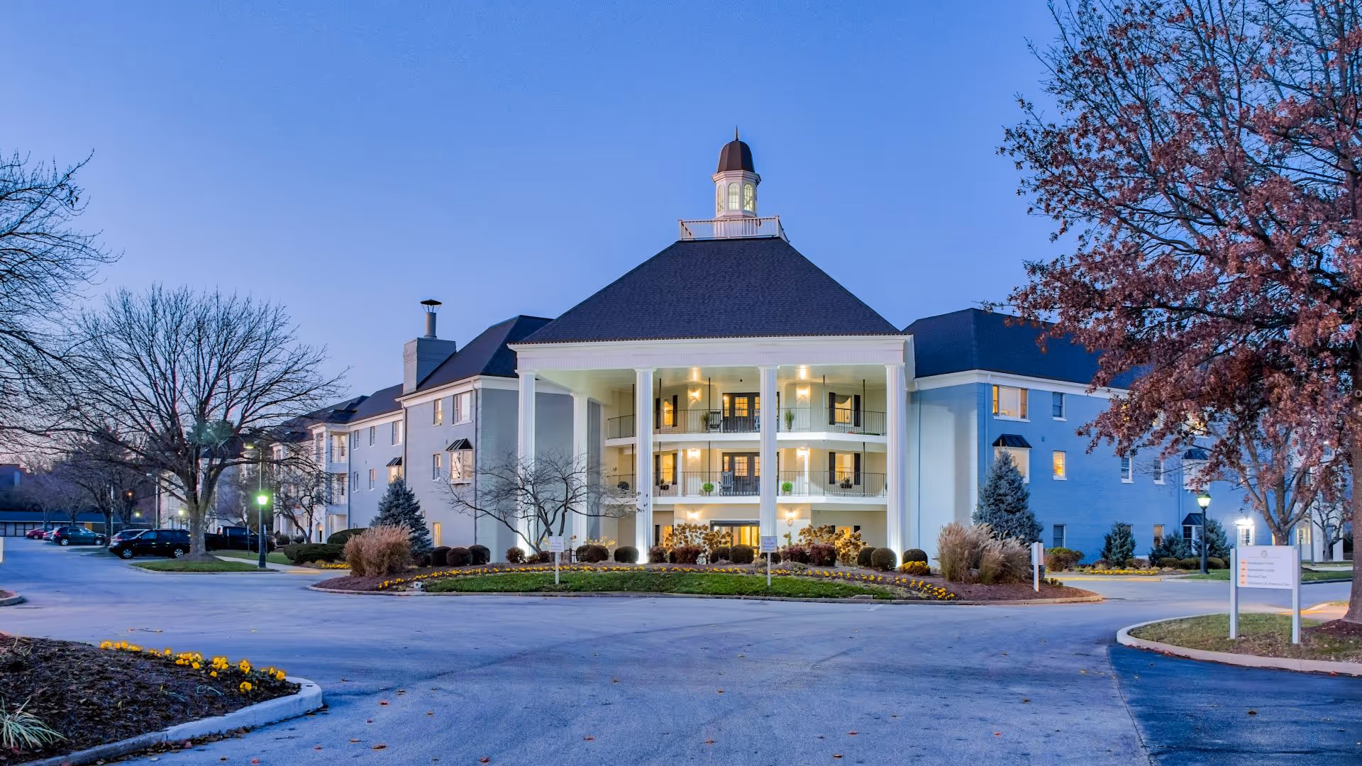 Front exterior of a multi-story senior living building with large white columns, balconies, and a landscaped circular driveway at dusk.