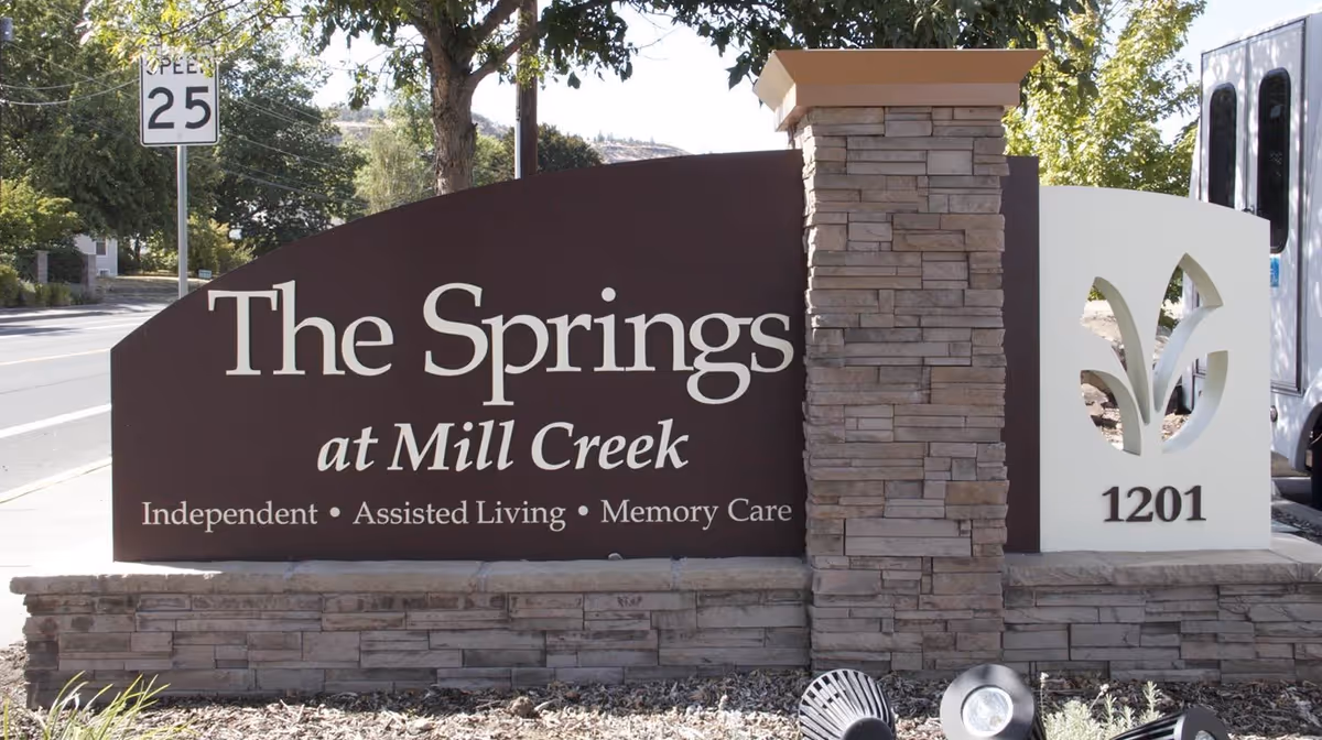 Entrance stone sign reading "The Springs at Mill Creek" with logo, address 1201 and surrounding landscaping.