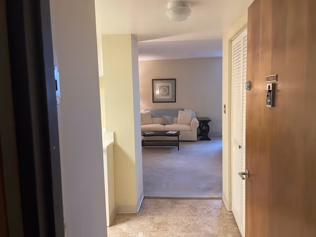 View from an entry hallway into a living room area with a beige sofa, a coffee table, and a side table with a framed picture on the wall above the sofa. The floor in the hallway is tiled, and the living room floor is carpeted.