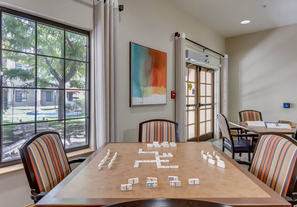 Bright common activity room with a table laid out for dominoes, striped chairs, a large window, and glass doors.