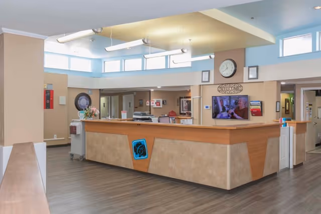 Reception area of Carrollton Health & Rehabilitation Center with a large wooden and beige front desk, a wall clock, a welcome sign, a mounted TV displaying a person, and overhead fluorescent lighting. The area has light-colored walls and windows near the ceiling allowing natural light.