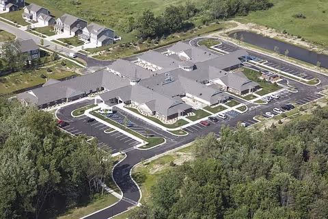 Aerial view of The Willows at Okemos: a single-story senior living complex with multiple wings, parking lots, surrounding lawns, a pond, and nearby houses.