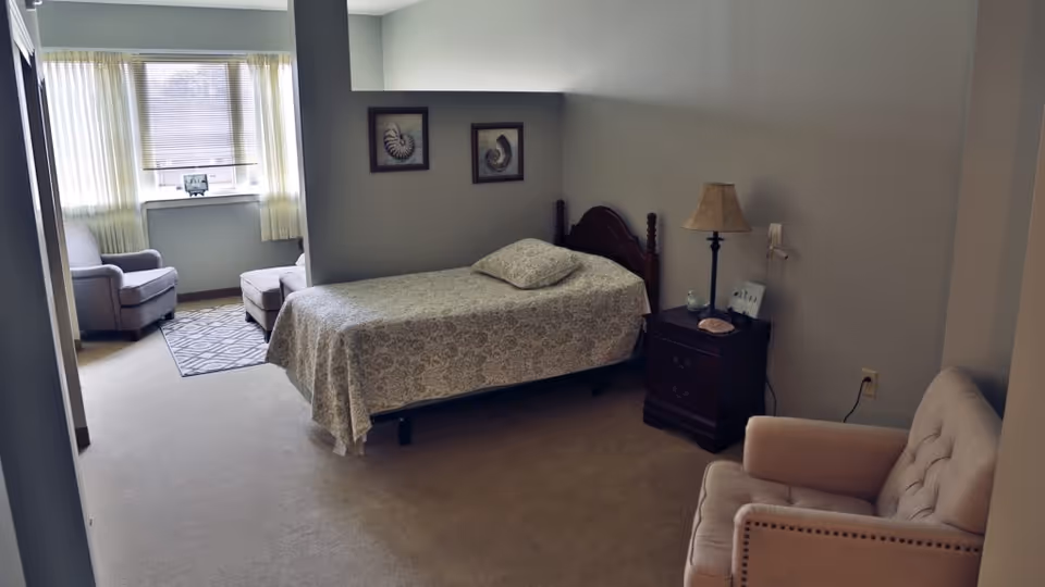A cozy bedroom in a senior living facility featuring a single bed with a patterned bedspread and a pillow, a wooden nightstand with a lamp and decorative items, a beige armchair, and a sitting area near a window with two upholstered chairs and a small rug. The room has light-colored walls and carpeted flooring.
