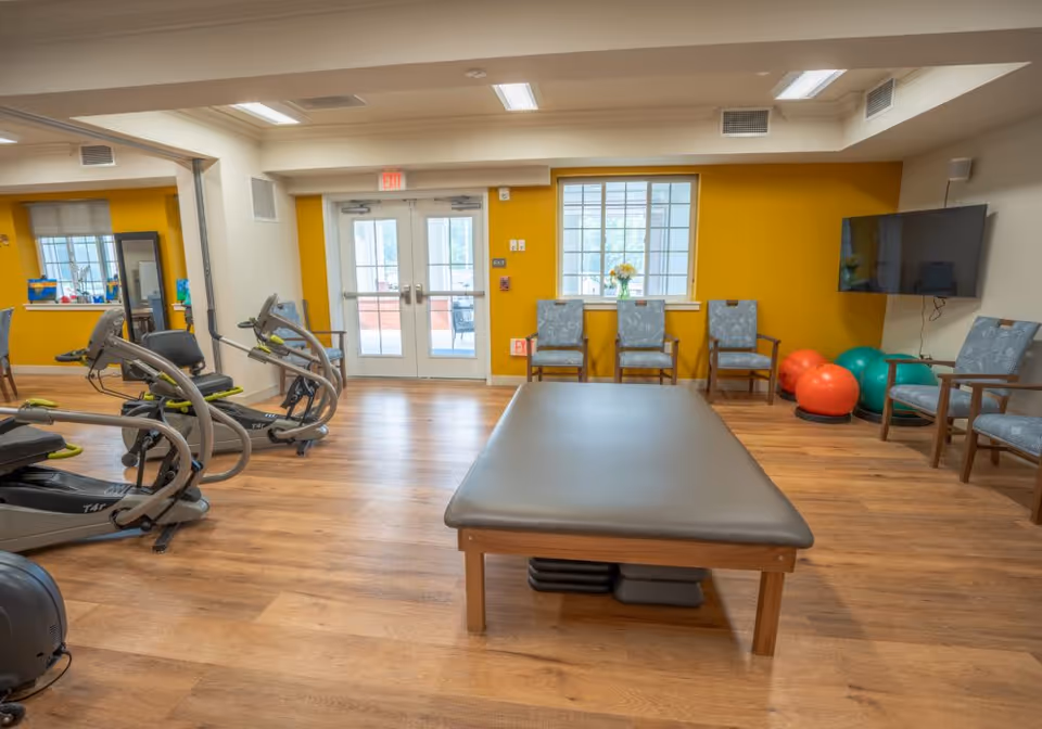 Bright therapy/fitness room with exercise equipment, a central treatment table, chairs and exercise balls.