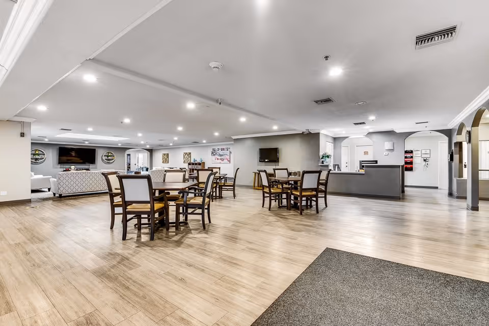 Spacious senior living common area with wooden flooring, multiple round tables with chairs, a reception desk, and a lounge area with sofas and a wall-mounted TV in the background.
