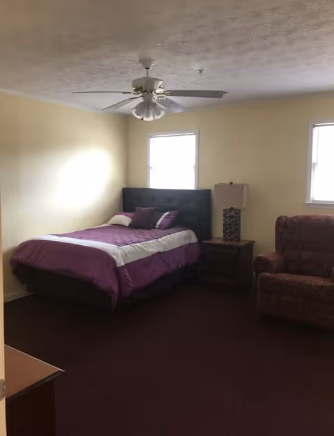 Bedroom with a purple-covered bed, nightstand and lamp, upholstered armchair, two small windows and a ceiling fan.