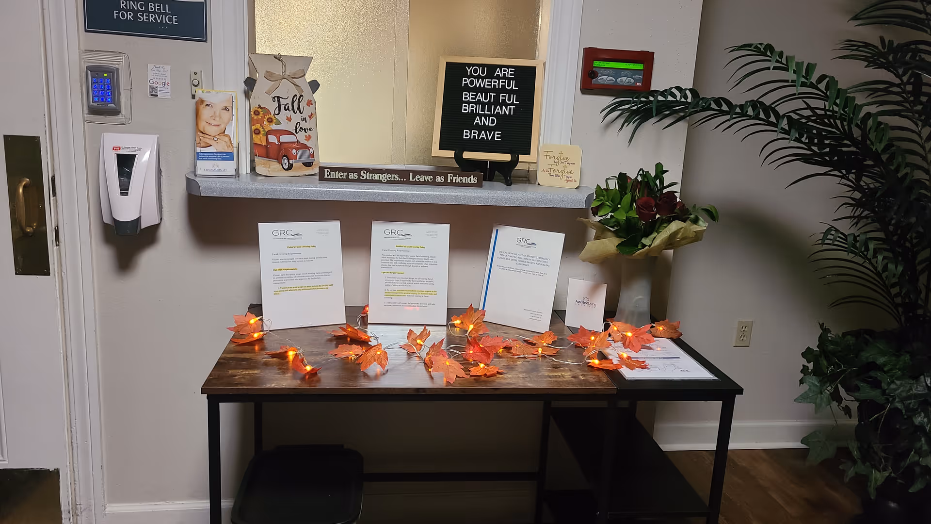 Reception area with a wooden table decorated with autumn leaf string lights, informational brochures, a bouquet of flowers, and a small sign that reads 'Enter as Strangers... Leave as Friends'. A letter board on the counter behind the table displays the message 'YOU ARE POWERFUL BEAUTIFUL BRILLIANT AND BRAVE'. A hand sanitizer dispenser is mounted on the wall next to a door with a sign that says 'RING BELL FOR SERVICE'.