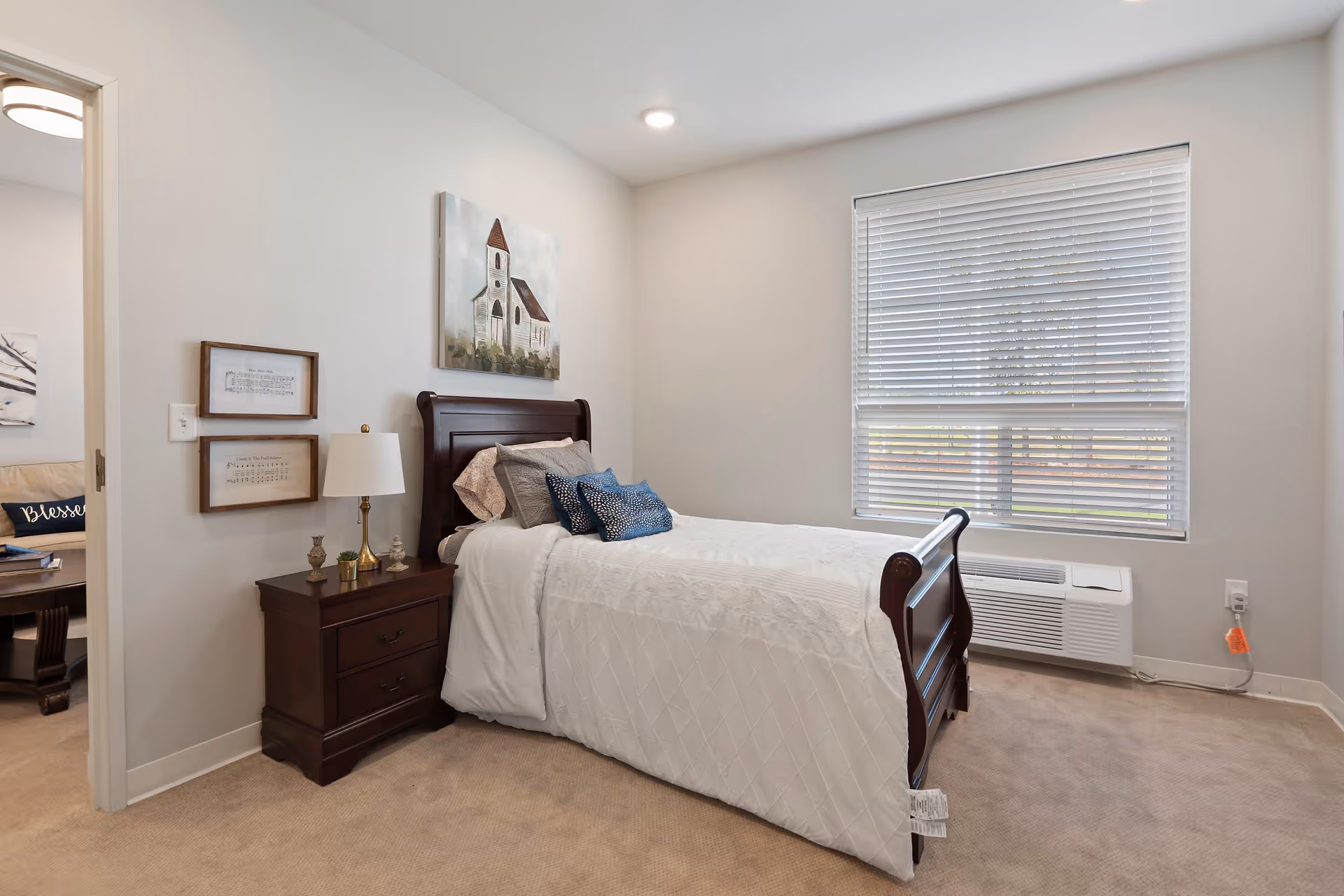 A neatly made single bed with white bedding and blue accent pillows in a bedroom. Next to the bed is a dark wooden nightstand with a lamp and decorative items. On the wall above the bed is a painting of a church. A window with white blinds lets in natural light. The room has beige carpet and light-colored walls. A doorway leads to another room with a couch and a pillow that says 'Blessed'.