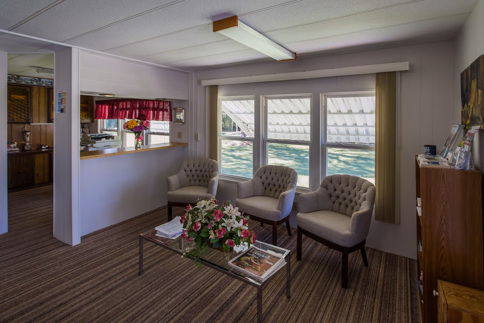 A cozy waiting area with three tufted armchairs arranged around a glass coffee table adorned with a floral arrangement and magazines. The room has large windows with vertical blinds partially open, allowing natural light to brighten the space. A wooden bookshelf with brochures and magazines is positioned against the wall, and a pass-through window with a red valance connects to an adjacent room.
