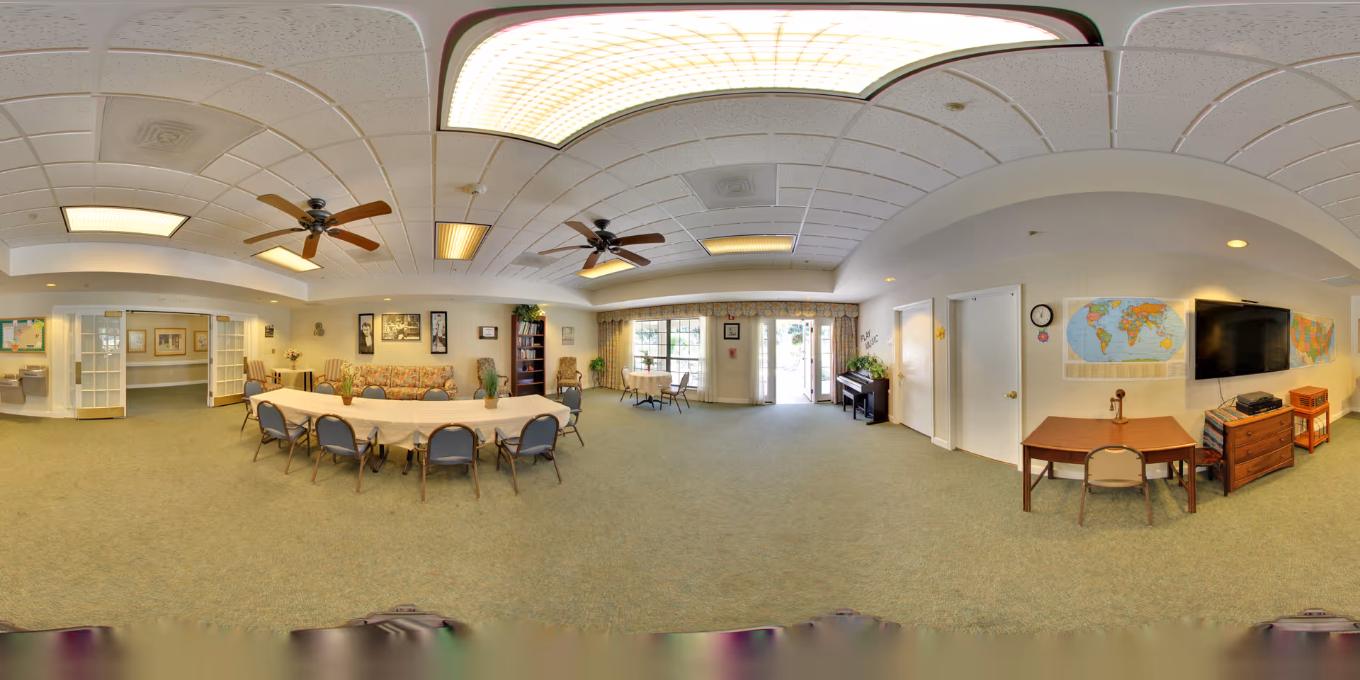 Spacious senior living community common room with a long dining/activity table and chairs, a seating area with sofa, TV, piano, and large windows.