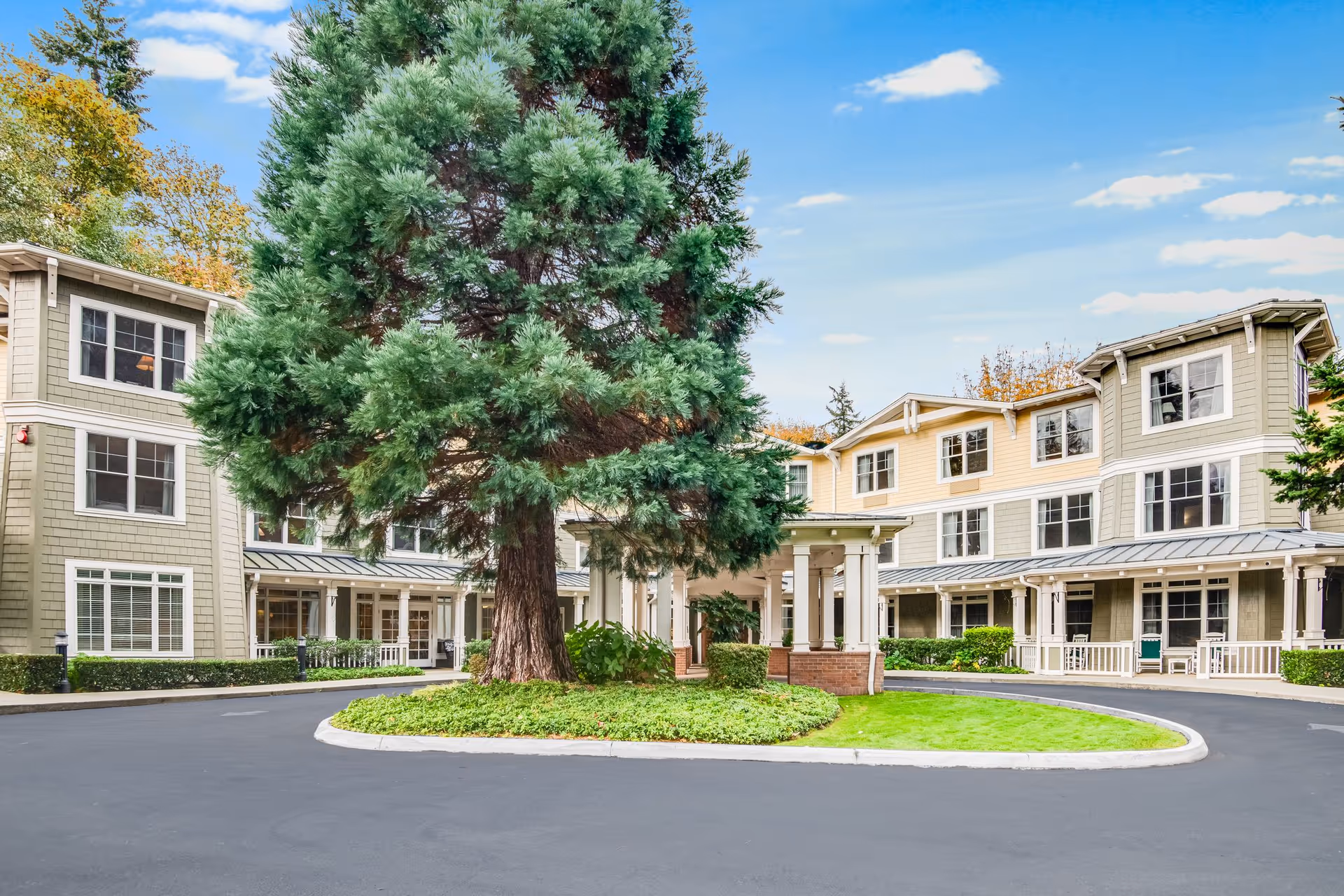 Exterior view of a senior living facility with a large tree in the center of a circular driveway. The building has multiple windows, a covered entrance, and a well-maintained lawn and shrubs under a blue sky with scattered clouds.