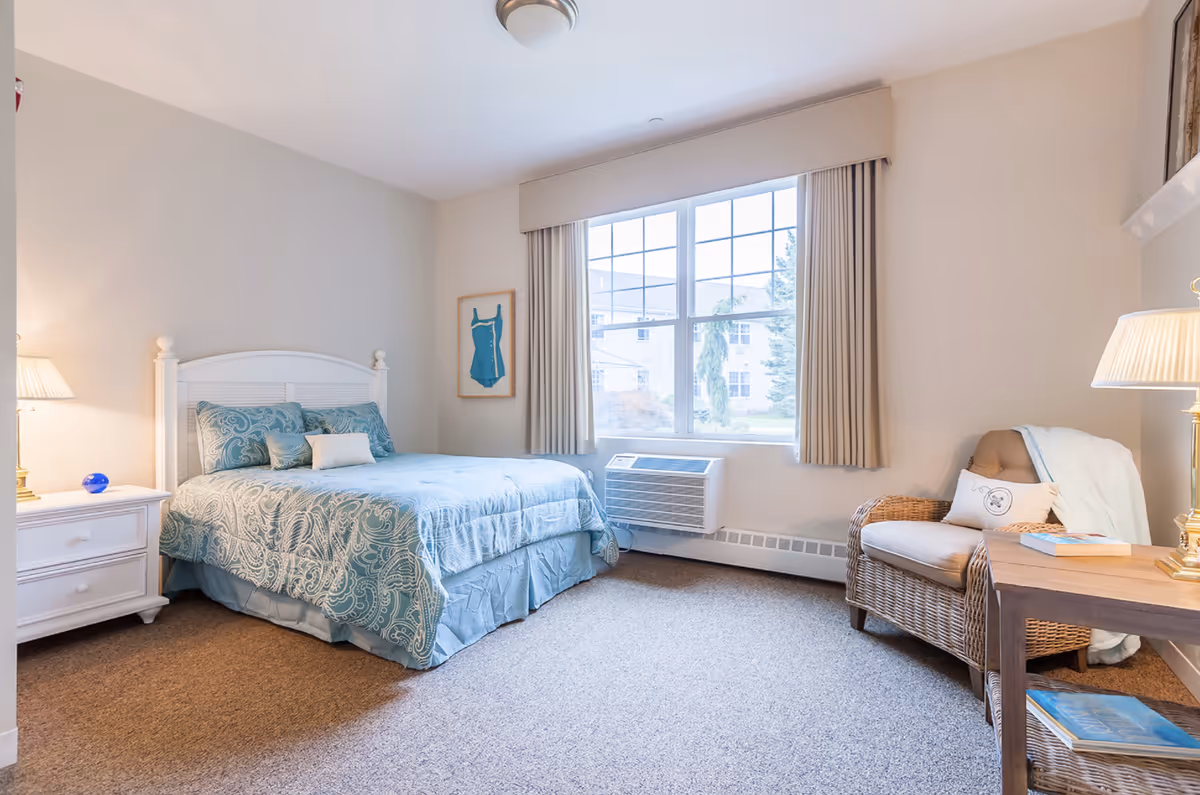 A bright and cozy bedroom with a large window letting in natural light. The room features a white wooden bed frame with blue patterned bedding, a white nightstand with a lamp and a small blue decorative item, a wicker armchair with a cushion and a light blue throw, and a wooden side table with books and a lamp. Beige curtains frame the window, and the carpet is a neutral tone.