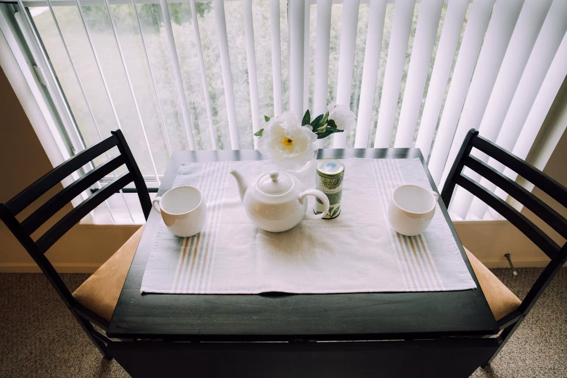 A small black dining table set for two with a white teapot, two white cups, a small container, and a white flower in a vase on a white placemat. The table is positioned in front of a window with vertical blinds.