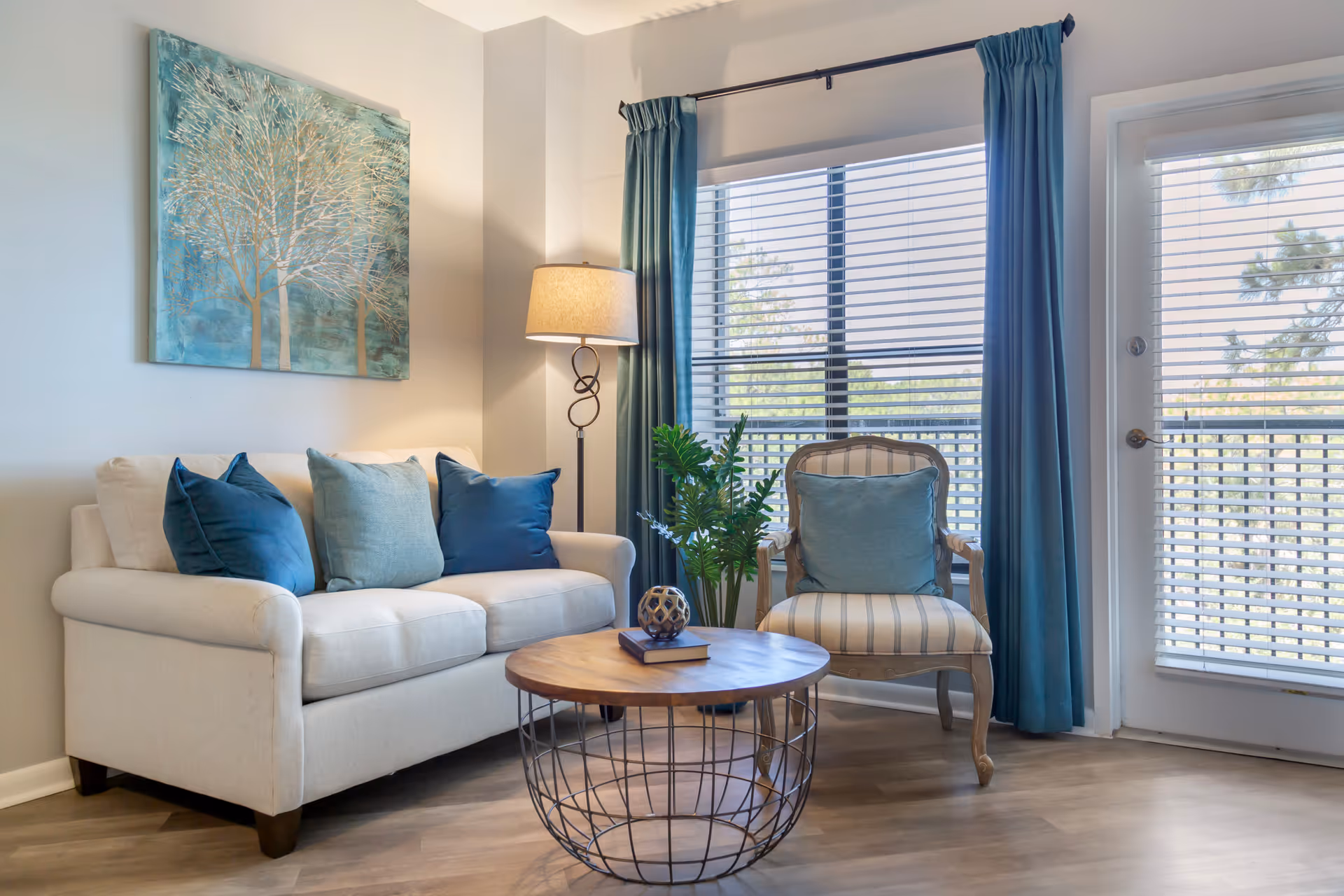 Bright living room with a cream sofa, striped armchair, round wooden coffee table, floor lamp, and large window with blue curtains.