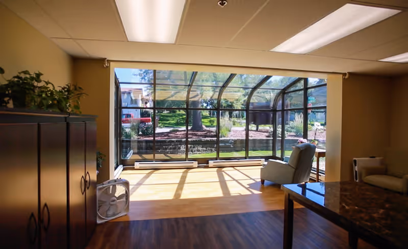 Sunlit seating area featuring a curved glass sunroom wall, chairs, a table and storage cabinets.