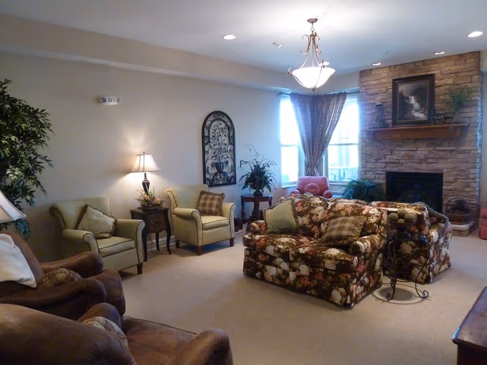 A cozy living room with floral patterned sofas, green armchairs, a stone fireplace with a framed picture above it, a chandelier, and large windows with curtains letting in natural light. There are decorative plants and a side table with a lamp.
