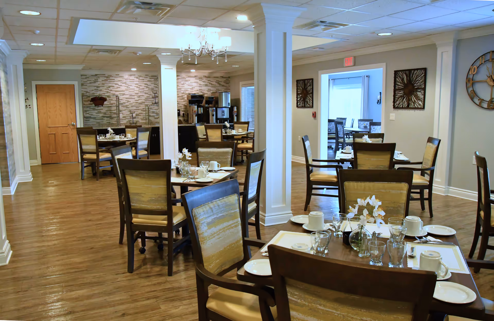Bright dining room with multiple tables set for meals, wooden chairs, and decorative wall accents.
